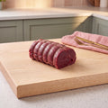 A single boned and rolled venison haunch joint, neatly tied at even intervals with natural butcher's twine, its deep ruby-red to dark crimson exterior showing the lean quality of the meat, sitting centrally on a clean pale ash chopping board in a warm, contemporary home kitchen with sage green cabinetry and warm natural light.