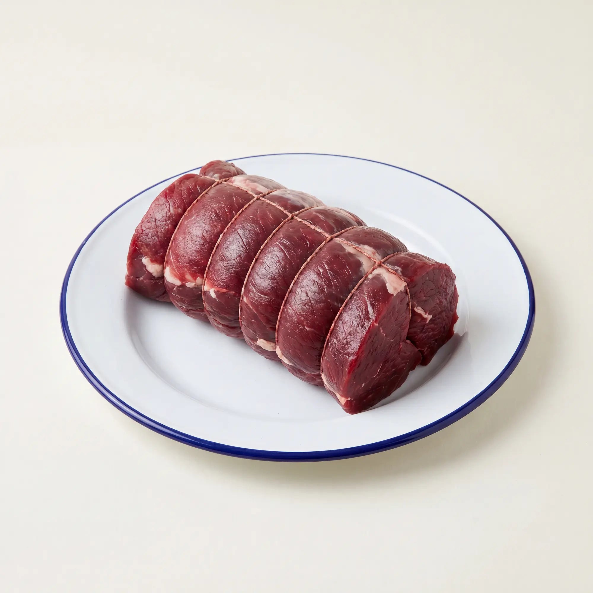 Raw venison haunch (boned and rolled) on a white plate with a blue rim on a light beige background