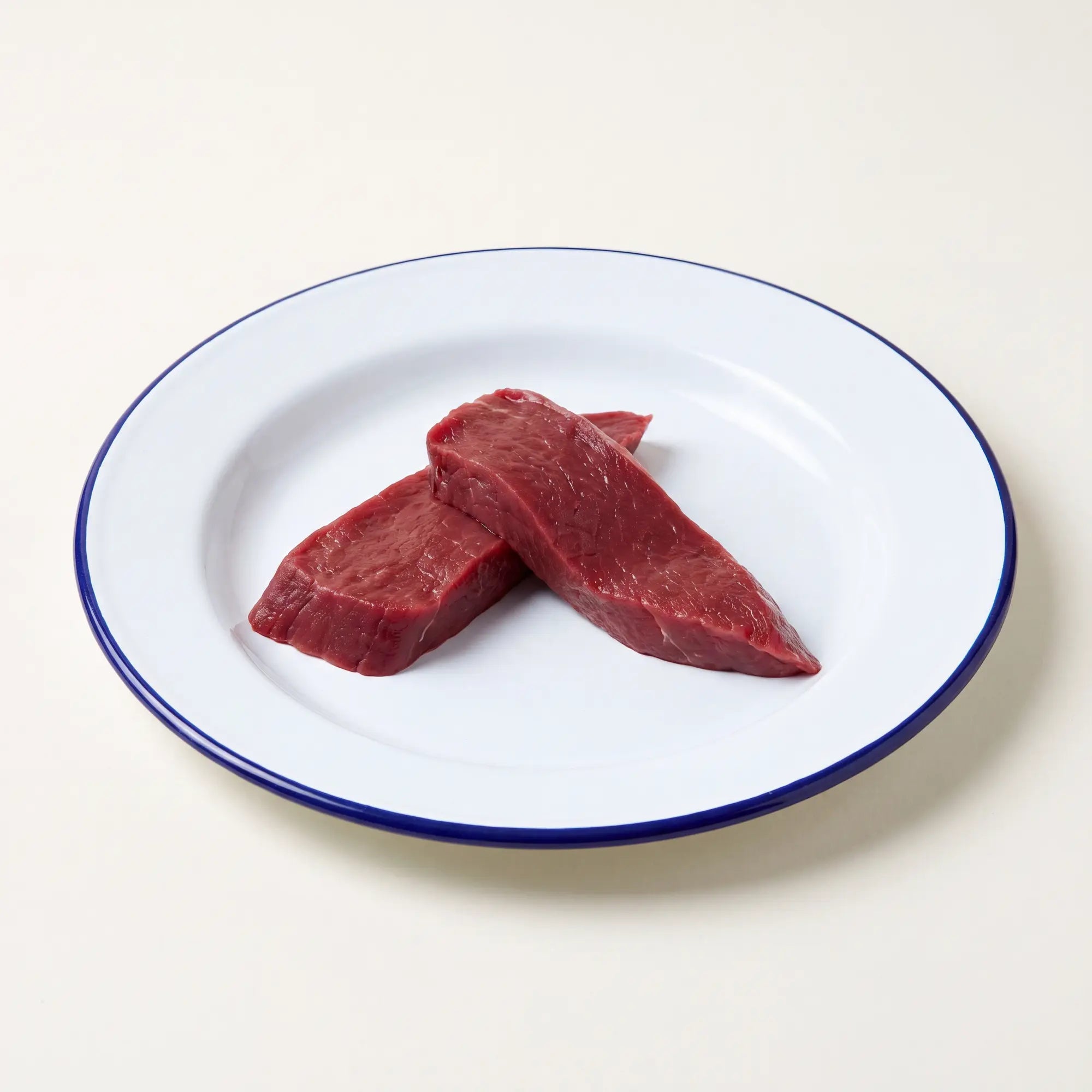 Two raw venison fillet steaks on a white plate with blue rim with a beige background.