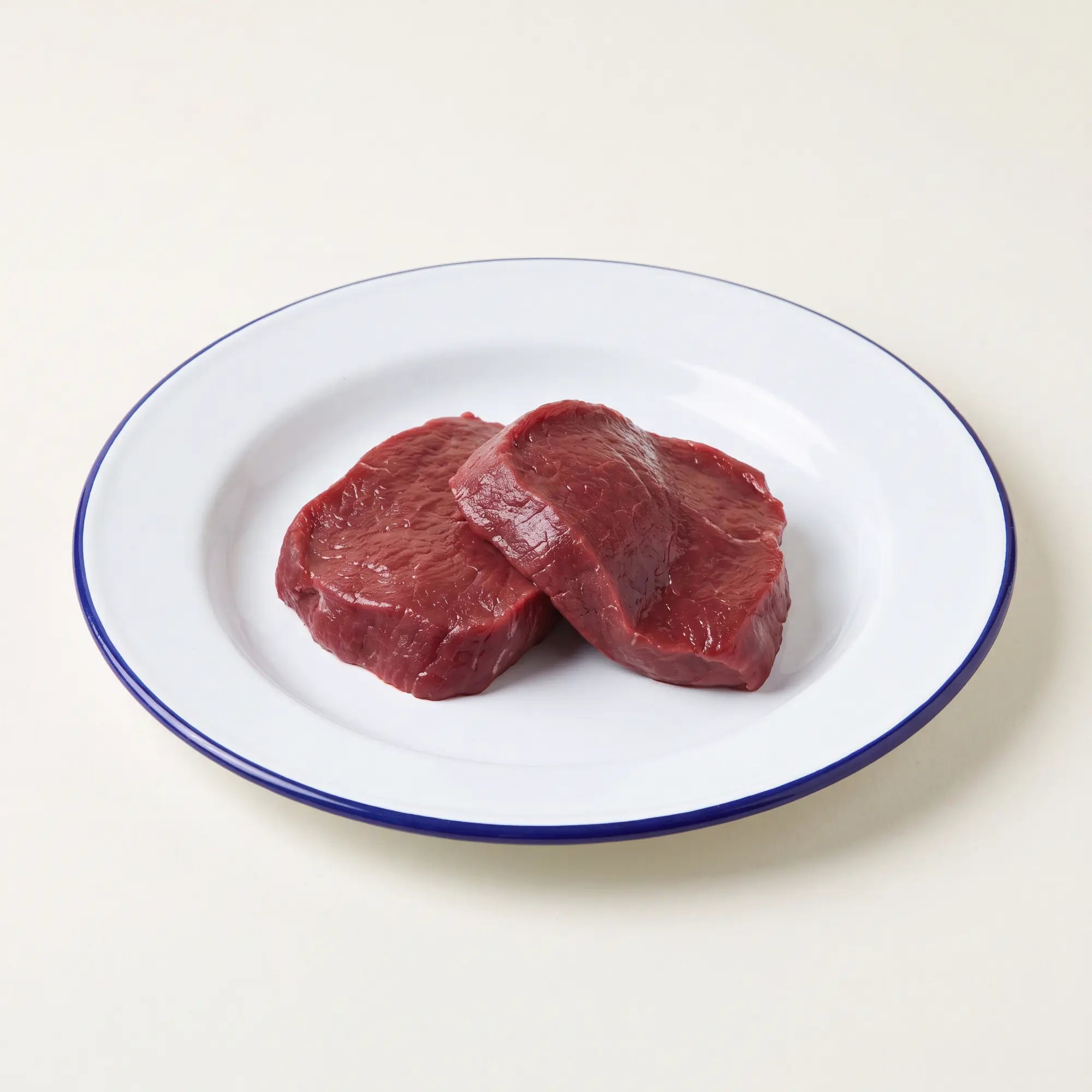 Two raw wild venison pave steaks on a white plate with a blue rim against a beige background