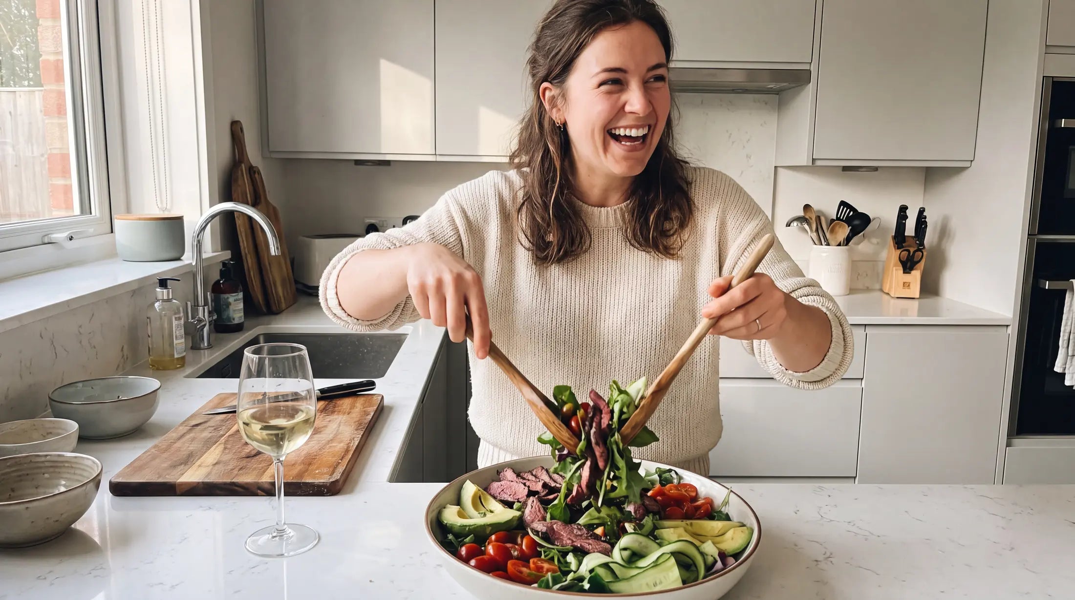 Woman laughing while tossing a fresh venison salad with avocado, cherry tomatoes and mixed leaves in a bright modern kitchen.
