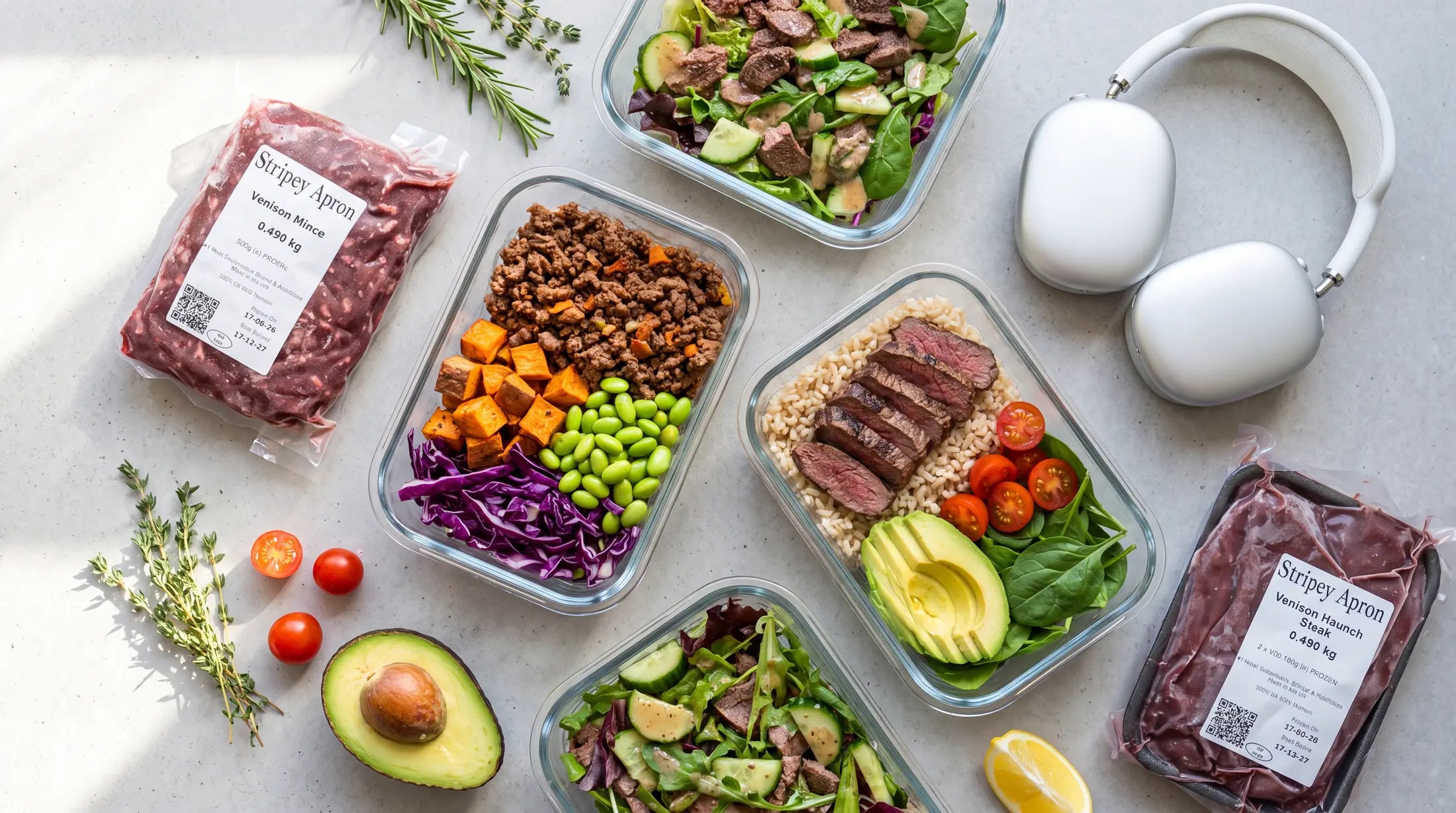 Overhead flat lay of colourful venison meal prep containers with Stripey Apron venison packets, sliced avocado, sweet potato, edamame and fresh herbs on a light marble surface, with silver headphones.